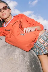 Bolso de nylon color naranja quemado, con textura sutil y manija fruncida. Presenta un parche rectangular negro con el logo 'KUL.' en blanco en el frente.