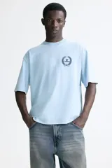 Remera de algodón color celeste, con cuello redondo, mangas cortas y corte holgado. Presenta un pequeño estampado frontal en el pecho con diseño de corona de laureles y texto.