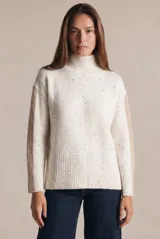 Polera de punto con diseño oversize, color blanco con motas multicolores. Presenta cuello alto y paneles laterales en las mangas con textura acanalada en tono beige.