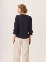 Remera de manga larga color negro con cuello en V y puños fruncidos. Presenta un estampado sutil en tonos grises y blancos en el centro del pecho, simulando un ramillete de flores secas.