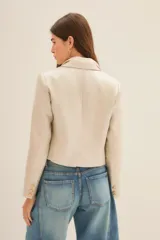 Saco corto tipo blazer en color beige, confeccionado en lino con acabado brillante. Presenta solapas clásicas, cierre frontal con un botón y mangas largas con puños abotonados.