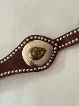 Cinto faja de cuero marrón con tachas metálicas plateadas en los bordes. Presenta una pieza central metálica con textura martillada y un dije en forma de gota con piedra natural.