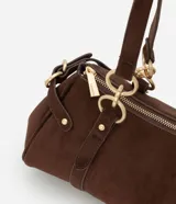 Cartera mediana de diseño alargado y estructura firme, confeccionada en gamuza sintética de color marrón. Cuenta con doble asa fina sujeta por argollas metálicas doradas, cierre superior y detalles de solapas decorativas con tachas en los laterales.