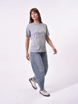 Remera de manga corta color beige claro con cuello redondo. Presenta un estampado gráfico central con un plato de pasta rodeado por un borde azul y blanco, sobre un fondo de franjas verticales rojas y blancas. El estampado incluye texto en cursiva en tonos turquesa y rojo.