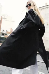 Trench largo de color negro con cuello de solapa, cierre frontal cruzado con botones y cinturón ajustable a tono. Presenta mangas largas con trabillas en los puños, bolsillos delanteros de vivo y abertura en la espalda.