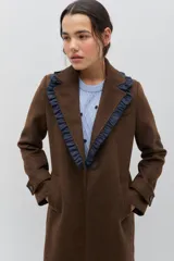 Tapado de lana color marrón con silueta entallada. Presenta solapas clásicas decoradas con un volado en contraste de color azul oscuro, cierre frontal con un botón y bolsillos laterales. Las mangas cuentan con detalles de botones en los puños.