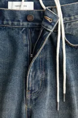 Jean de cinco bolsillos en denim de algodón rígido, color azul oscuro con efecto desgastado. Modelo de cintura clásica con cierre y botón, corte holgado y piernas anchas tipo barrel con costuras laterales marcadas.