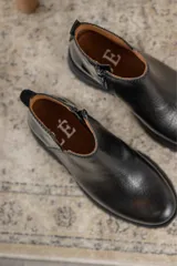 Botinetas negras con suela track, combinando cuero texturizado en la parte frontal y cuero charolado en la parte trasera. Diseño de caña baja con detalle de corte lateral.