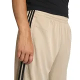 Pantalón de jogging Adidas de corte regular en color beige, con las tres tiras características en negro a lo largo de los laterales y el logo del trifolio bordado en la parte frontal.
