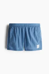 Short de baño de nylon en color azul, con cintura elástica y cordón de ajuste interno. Presenta bolsillos laterales, un bolsillo trasero con cierre autoadherente y un pequeño parche con logo en la parte inferior. Incluye suspensor interno de malla.