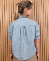 Camisa de denim de algodón celeste, con cierre frontal de botones, cuello alto con volados y mangas cortas abullonadas con puños elastizados fruncidos.