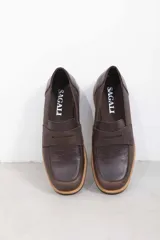 Mocasín de cuero vacuno en color marrón, con diseño bitono que combina texturas lisas y con grabado tipo cocodrilo en el empeine. Cuenta con detalle de estribo clásico y suela plana con vira en contraste.