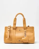 Cartera tipo tote XL confeccionada en símil cuero de textura granulada color habano. Cuenta con doble asa de mano, detalles de correas decorativas con hebilla en los laterales y un charm colgante en forma de corazón. Incluye placa metálica con las iniciales de la marca en el frente.