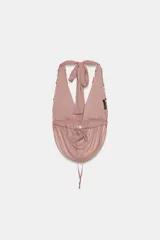 Top halter de color rosado con escote en pico pronunciado, confeccionado en tejido semitransparente con efecto arrugado y detalle de lentejuelas brillantes en la parte superior. Cuenta con tirantes para anudar al cuello y espalda descubierta con lazo ajustable.