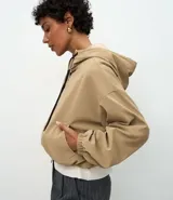 Campera cortavientos de color beige con diseño balonê, capucha con cordones ajustables, cierre frontal de cremallera y puños con elástico. Presenta un corte holgado y moderno.