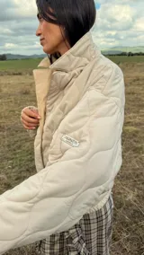 Campera puffer color beige con diseño acolchado en rombos, cuello alto, cierre frontal y solapa triangular decorativa con iniciales bordadas.