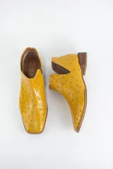 Botin tipo Chelsea de cuero con grabado efecto avestruz, color amarillo mostaza. Presenta tajos laterales, puntera cuadrada y taco bajo de madera.