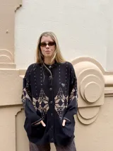 Saco tejido de punto con diseño geométrico en tonos negro y beige. Presenta cierre frontal con botones, cuello redondo, mangas largas y dos bolsillos delanteros. Incluye detalles bordados en el pecho.