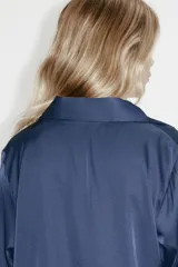 Blusa de tela satinada con brillo, cuello clásico, escote en V con cierre frontal de botones, mangas largas con puños sencillos y bajo redondeado.
