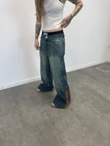 Jean de corte amplio con lavado desgastado en tono azul verdoso. Presenta bordados lineales en ambas piernas y cierres laterales metálicos que, al abrirse, revelan un panel de pana en color marrón. Cuenta con bolsillos amplios y botones personalizados.