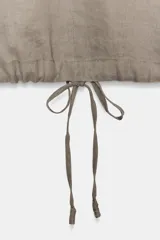 Top de lino de color beige, con cuello redondo, manga corta, bolsillos frontales aplicados y bajo ajustable con cordones. Cuenta con abertura en la espalda y cierre de botón.