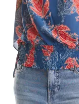 Blusa de gasa con estampado de arabescos en tonos azules y rojos, con cuello a la base y mangas tipo capa. Presenta una silueta holgada con ruedo elástico en punto smock que ajusta a la cintura.