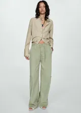 Camisa color beige de corte crop, confeccionada en mezcla de lino y lyocell. Tiene cuello camisero, mangas largas con puños abotonados, lazo ajustable en la cintura y cierre frontal con botones.