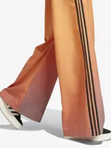 Pantalón deportivo Adidas de corte holgado con diseño degradado en tonos naranja y marrón. Presenta las tres rayas negras características de la marca en los laterales y logo bordado en la parte superior.