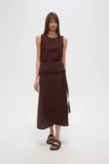 Falda larga de algodón color marrón oscuro, de corte envolvente (wrap skirt) con abertura lateral y borde deshilachado. Se ajusta con un lazo lateral que pasa por argollas metálicas.