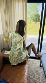 Camisa larga tipo sobrecamisa o abrigo ligero, color verde menta pálido con estampado floral en tono amarillo claro. Presenta escote en V con atado frontal mediante lazos, mangas largas con detalle fruncido y puños con flecos en tono turquesa.