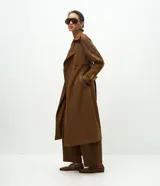 Trench coat largo de gamuza sintética color marrón oscuro, con cuello solapa, cierre frontal cruzado con botones y cinturón ajustable en la cintura. Presenta mangas largas con reguladores en los puños y forro interior.
