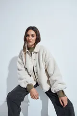 Campera de corderito color beige, con cuello camisero y cierre frontal de botones a presión. Presenta una cartera central lisa en contraste y mangas largas con puños elásticos.