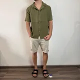Camisa de hombre color verde oliva, de muselina con textura arrugada, manga corta, corte oversize, cuello cubano y cierre con botones. Presenta un pequeño logo bordado en el pecho.