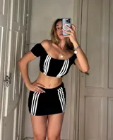 Conjunto deportivo de dos piezas color negro con tres bandas blancas verticales. El top es de escote hombros descubiertos y mangas cortas, mientras que la minifalda presenta un diseño ajustado con las mismas bandas laterales.