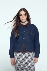 Campera de jean color negro con cuello de pana en tono marrón, cierre frontal con botones y bolsillos en el pecho. Presenta un diseño de corte relajado y ligeramente cropped, con interior forrado en corderito para mayor abrigo.