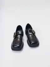 Zapato tipo guillermina de cuero negro con punta cuadrada y suela de plataforma. Presenta una correa ajustable sobre el empeine con hebilla metálica y un panel frontal con costuras decorativas en contraste.