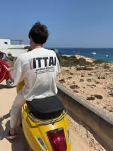 Remera marrón de corte holgado con estampado en la espalda que incluye la palabra "ITTAI" en blanco con líneas rojas y azules, la frase "AN IBIZA PRODUCE" y un texto más pequeño que dice "Ittai Stars Department Gallery - Ibiza Edition. If you're in Ibiza, 112 is not an option.".