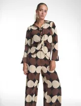 Conjunto de dos piezas compuesto por camisa de manga larga con nudo frontal y pantalón estilo palazzo de pierna ancha. Ambas prendas presentan un estampado geométrico de círculos grandes en tonos marrón, negro y beige. La camisa cuenta con cuello solapa y cierre frontal con botones, mientras que el pantalón posee tiro alto y corte holgado.