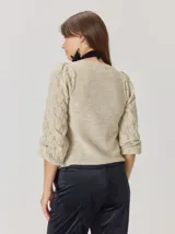 Buzo de lana color beige con mangas abullonadas de tejido calado con diseño de rombos. Presenta un cuello redondo y un detalle de flor negra de tela con cintas colgantes en el escote.