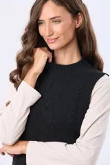 Chaleco tejido de punto grueso en color beige, con cuello redondo y diseño estructurado con detalles de trenzas verticales. Presenta un pequeño aplique de cuero sintético en la parte inferior.