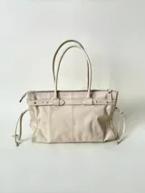 Bolso grande de cuero marrón con textura tipo cocodrilo, estructura rígida y asas para llevar al hombro. Tiene una tira con tachas y hebillas en cada extremo, también lleva lazos en los costados.