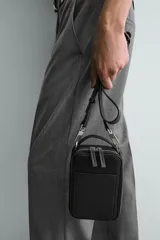 Bandolera negra con textura grabada, forma rectangular y cierre de cremallera. Tiene un bolsillo interior plano y otro frontal plano. Incluye asa de hombro ajustable y extraíble.