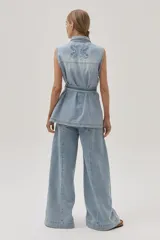 Chaleco largo de jean celeste con cuello camisero, sin mangas, con bolsillos frontales, laterales e internos, y lazo ajustable en la cintura.