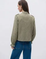 Camisa de manga larga con estampado de cuadros vichy en tonos verde oliva y crudo. Presenta cuello mao, cierre frontal con botones y volados decorativos en la parte delantera. Las mangas terminan en puños elastizados con un ligero acabado de volado.