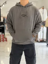 Canguro oversize beige con capucha y cordones ajustables, bolsillo tipo canguro y pequeño bordado en el pecho.