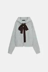 Chaqueta de punto gris con capucha, manga larga y cierre frontal con cremallera. Presenta un lazo decorativo de color marrón oscuro en contraste en la zona del cuello.