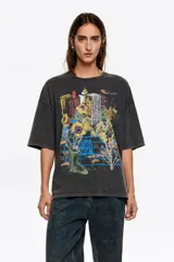 Remera de algodón, color gris oscuro con efecto gastado (acid wash), corte oversize y escote redondo. Presenta un estampado frontal artístico con una ciudad, flores amarillas y una bota de lluvia verde.