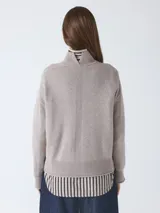Suéter de cuello alto, color beige topo, de manga larga y calce holgado. Presenta un borde de punto acanalado en el cuello, puños y bajo, con una camisa de rayas negras y beige asomando por el bajo.