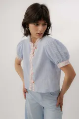 Blusa celeste a rayas blancas, de manga corta abullonada. Cuenta con cuello mao y tapeta de botones con detalles de estampado floreado en tono rosado, presentes también en el cuello y los puños.