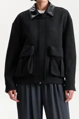 Chaqueta tipo bomber de corte amplio en color negro con textura, cuello camisero en contraste de material sintético y grandes bolsillos frontales con solapa. Presenta un cierre frontal oculto por una solapa.